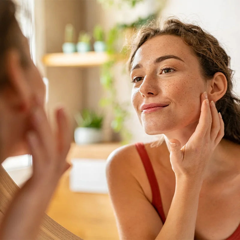 A young woman with clear, glowing skin and freckles smiles softly while gently touching her cheek as she looks in the mirror, reflecting post-treatment skin confidence and healthy radiance.